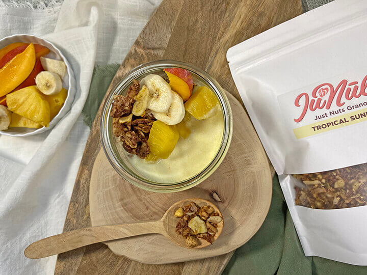 Photo of a bag of Tropical Sunrise JuNūla Just Nuts Granola on a table next to a bowl of granola with yogurt and fresh fruit