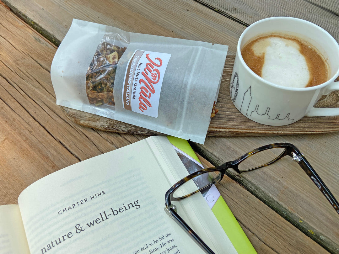 Photo of a 2oz bag of Cappuccino Italiano JuNūla Just Nuts Granola on a table next to an open book, some reading glasses, and a cup of cappuccino