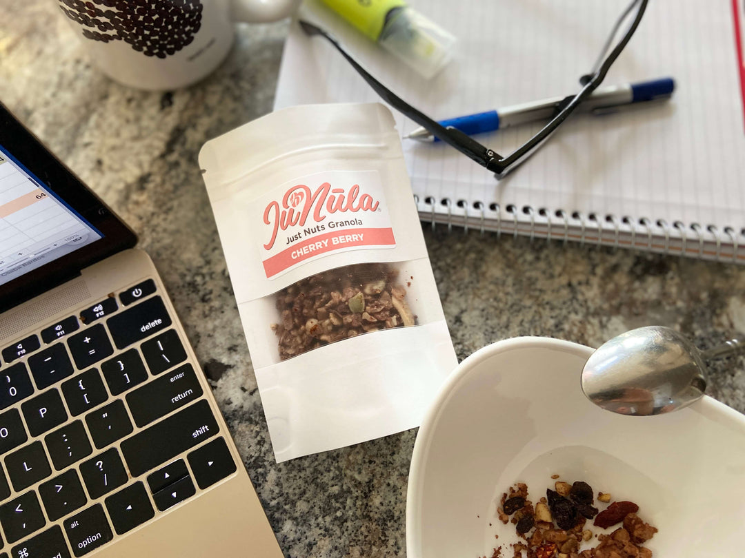 Photo of a 2oz bag of Cherry Berry JuNūla Just Nuts Granola on a desktop next to a laptop and a notebook with a pen